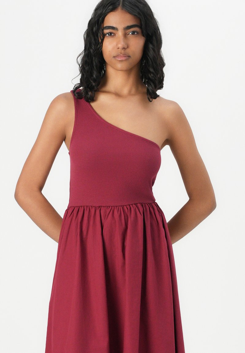 Robe bordeaux à une épaule avec un corsage ajusté en maille et une jupe évasée et froncée. Texture lisse, silhouette décontractée, sans motifs visibles.