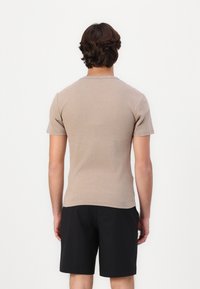 Beige t-paita lyhythihaisella ja pyöreällä kaula-aukolla, slim fit -mallia ja sileää tekstuuria, yhdistettynä mustiin shortseihin. Takana näkyy vähäistä yksityiskohtaisuutta.