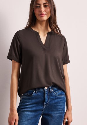 Femme portant un chemisier marron foncé à manches courtes avec une fente en V, rentré dans un jean en denim bleu, debout devant un fond uni.