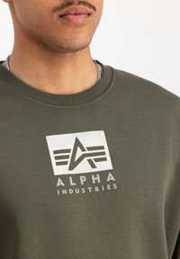 Olivovo zelená mikina s rebrovaným výstrihom, obsahujúca biely logotyp so stylizovaným "A" a textom "ALPHA INDUSTRIES" nižšie.