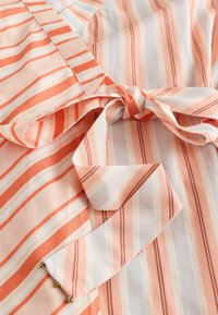 Tissu à rayures verticales et diagonales corail, pêche et blanches noué en nœud avec bords cousus visibles et petites décorations dorées.