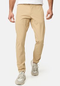 Pantalons légers beige à coupe fuselée, avec une texture lisse et des coutures subtiles. Associés à des chaussures de sport grises.
