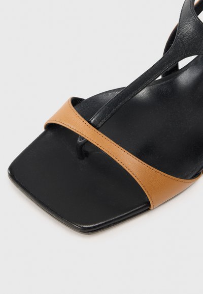 Sort sandal med firkantet tå, med en tan læderrem og en mat sort overdel. Designet inkluderer krydsremme med minimal hardware.