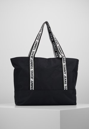 Duża czarna torba typu tote z białymi rączkami, na których znajduje się czarny napis "TOMMY JEANS", umieszczona na białym postumencie na tle jednolitego szarego tła.