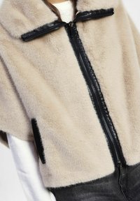 Primo piano di un gilet in pelliccia sintetica beige con finitura in pelle nera e zip, indossato sopra una maglietta a maniche lunghe bianca e jeans scuri.