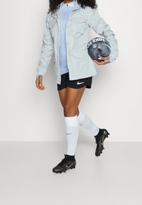 Ljusgrå jacka med flera fickor över en blå topp, svarta shorts, vita strumpor och svarta fotbollsskor; håller en grå fotboll.