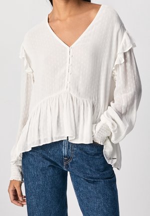 Kobieta w białej bluzce z dekoltem w kształcie litery V z guzikami, falbanami i marszczonymi mankietami, ubrana w niebieskie dżinsy, ręka w kieszeni.