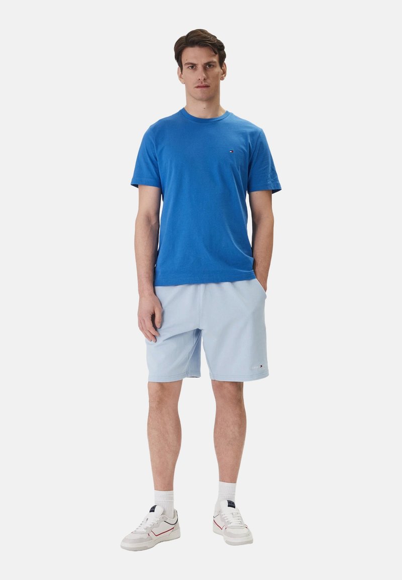 Homme debout portant un t-shirt à manches courtes bleu, un short bleu clair, des chaussettes blanches et des baskets blanches sur un fond uni.