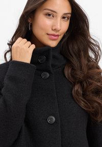 Black wool coat with a high collar, featuring three large black buttons and a soft, textured finish. Model holding the collar.