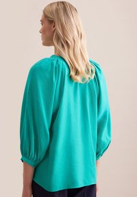 Blusa teal con maniche a sbuffo, texture liscia e dettagli plissettati sul retro. Leggermente più lunga sul retro con un orlo arrotondato.