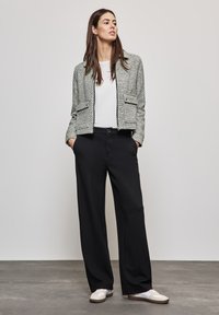 Gestructureerde lichtgrijze blazer met twee vo pockets, witte blouse, zwarte wijde pijpen broek en crèmekleurige sneakers op een grijze vloer.