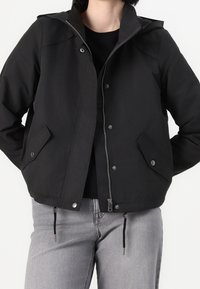 Czarna kurtka przeciwwiatrowa z zamkiem z przodu, bocznymi kieszeniami, elastycznymi sznurkami u dołu i dopasowanym kapturem. Gładka, matowa faktura.