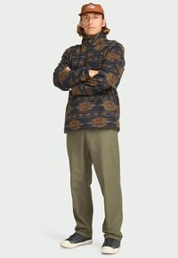 Man wearing brown patterned fleece pullover, olive green pants, brown cap, and black sneakers standing with arms crossed.