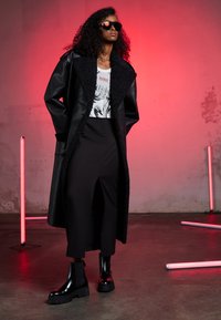 Palton din piele ecologică neagră cu guler texturat, tricou alb grafic, pantaloni negri cu talie înaltă și cizme negre cu platformă strălucitoare. Iluminare roșie.
