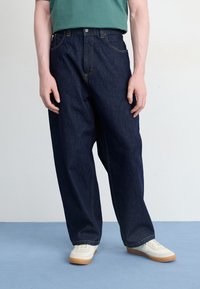 Temno modri jeans hlače z ohlapnim krojem, z rumenim šivanjem, sprednjimi žepi in gumbom za zapiranje. V kombinaciji z belimi supergami.
