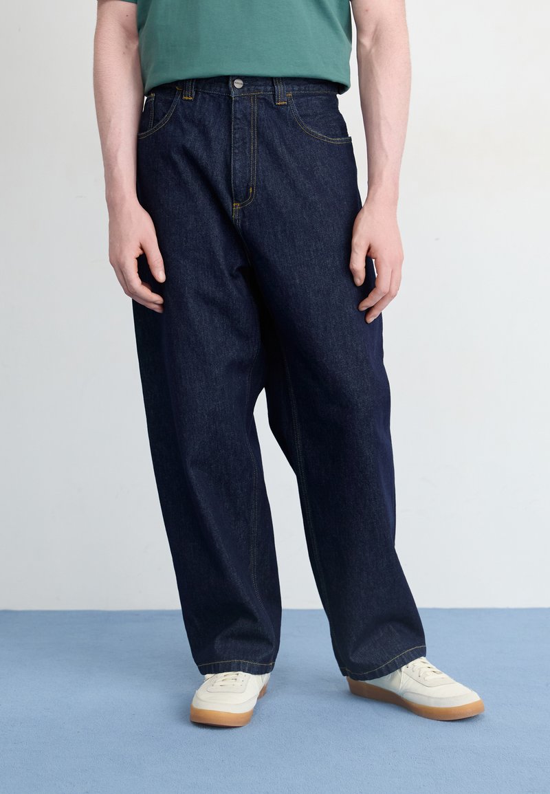 Temno modri jeans hlače z ohlapnim krojem, z rumenim šivanjem, sprednjimi žepi in gumbom za zapiranje. V kombinaciji z belimi supergami.