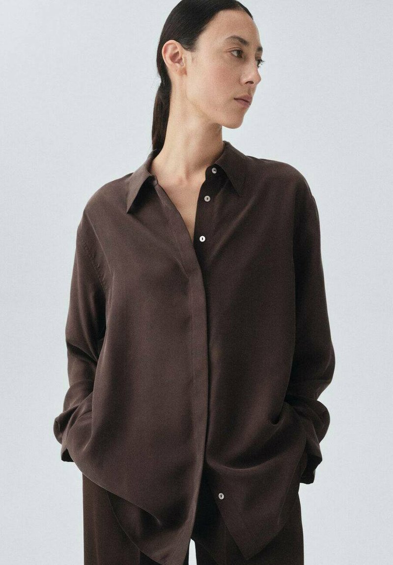 Camicia in seta marrone con colletto a punta, maniche lunghe e vestibilità rilassata. I dettagli includono piccoli bottoni bianchi e una texture liscia.