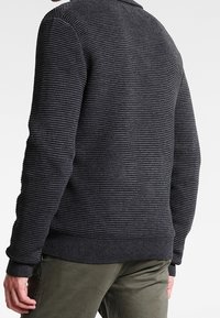 Mężczyzna w ciemnoszarej swetrze z prążkami i oliwkowych spodniach, widoczny od tyłu na białym tle.