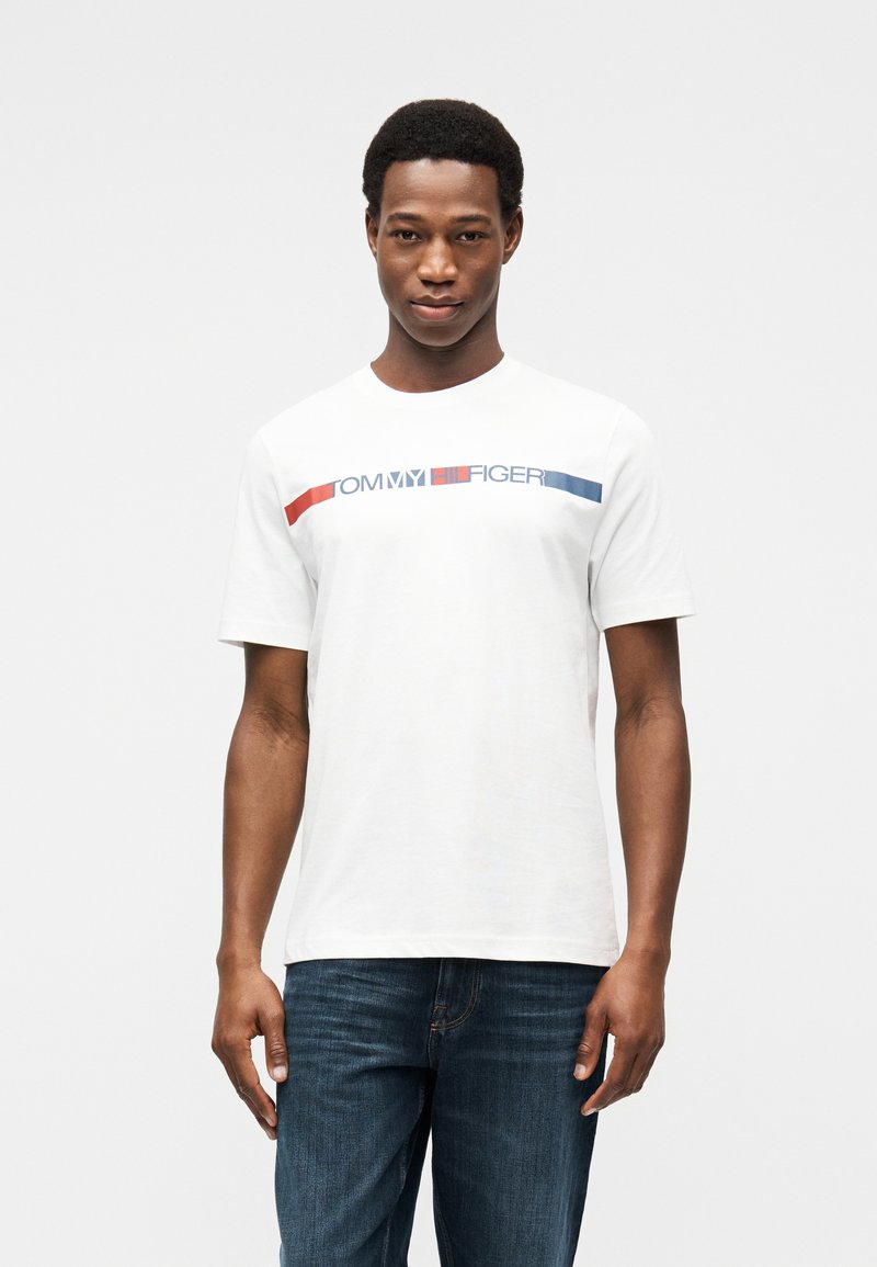 Λευκό βαμβακερό μπλουζάκι με στρογγυλή λαιμόκοψη, με γραφικό λογότυπο σε μπλε και κόκκινο χρώμα στο στήθος. Συνδυασμένο με μπλε τζιν παντελόνι.