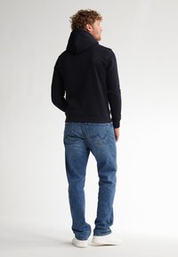 Sudadera negra con capucha ajustable, combinada con jeans de un azul claro y zapatillas blancas. El tejido parece suave; el corte es relajado con puños de canalé.