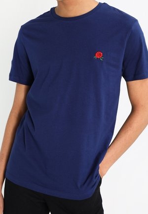 Osoba nosząca granatowy t-shirt z krótkim rękawem z małym czerwonym haftem róży na piersi, w połączeniu z czarnymi spodniami.