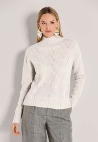 Frau, die einen weißen strukturierten Strickpullover mit Stehkragen, graue Karohosen und große geometrische Ohrringe aus Silber und Gold trägt.