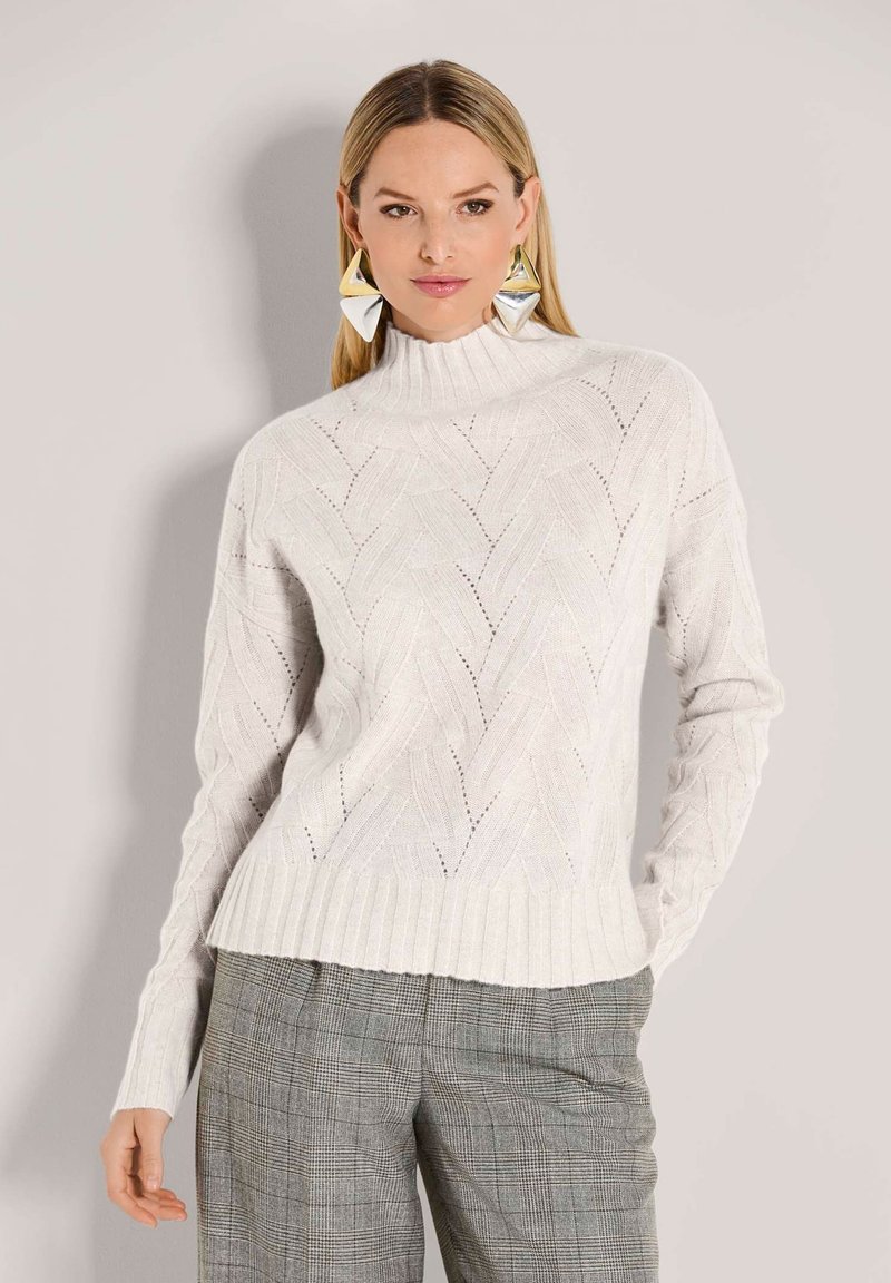 Frau, die einen weißen strukturierten Strickpullover mit Stehkragen, graue Karohosen und große geometrische Ohrringe aus Silber und Gold trägt.