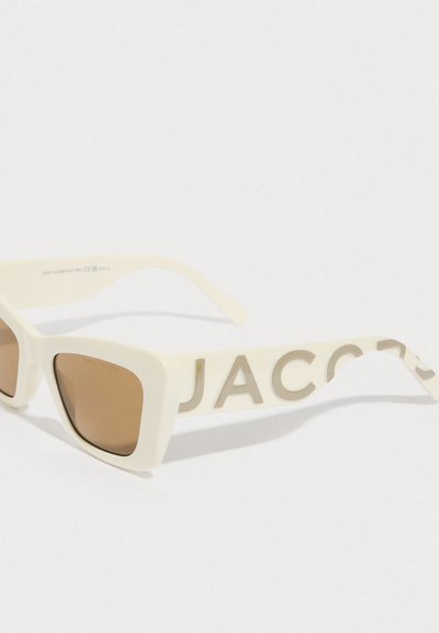 Unas gafas de sol rectangulares de color crema con lentes marrones y un gran texto "JACOBS" en las gruesas patillas, descansando sobre una superficie blanca.