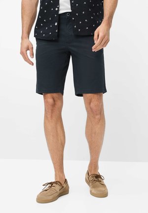 Mand iført navyblå shorts, sort kortærmet skjorte med tryk og beige snørebånds casual sko, stående på hvidt gulv.
