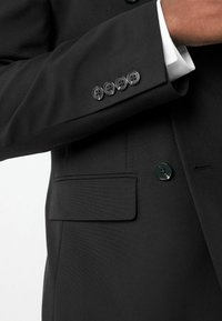 Chaqueta negra de traje con un bolsillo en el pecho, cinco botones negros en los puños y un botón verde en el cuello. Textura suave.