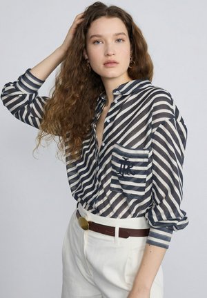 Femme aux longs cheveux bouclés portant une chemise boutonnée rayée bleu marine et blanc avec un logo sur la poche, pantalon blanc et ceinture marron, posant.