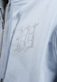 Vaaleansininen vetoketjullinen huppari, valmistettu pehmeästä kankaasta. Rintakehässä on koristeellinen hopeinen kristallilogo ja vetoketjussa teksturoitu vetoketjun vedin.