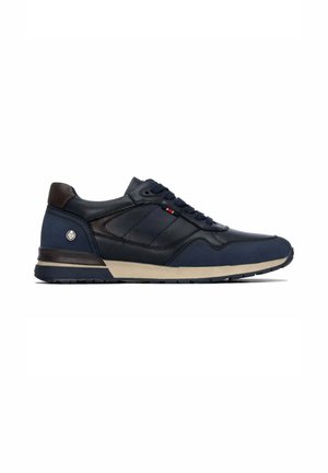 Zapatilla casual para hombre en azul marino con detalles negros, suela beige, cordones en la parte delantera y un pequeño logo metálico redondo cerca del talón.