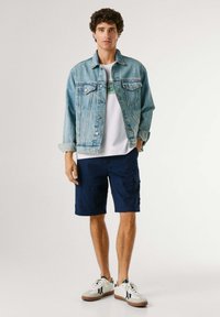 Noormees seisab, kandes helesinist teksatakki, valget graafilist T-särki, tumesiniseid cargo-lühikeseid pükse ja valgeid tossusid kummist tallaga.