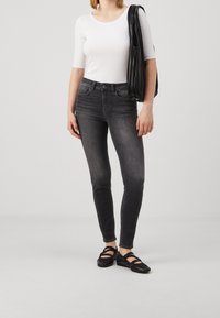 Pantalones vaqueros negros ajustados de cintura media, combinados con una blusa blanca de manga larga ceñida. Zapatos negros planos y un bolso negro plisado.