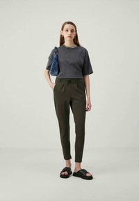 Jovem mulher a usar uma t-shirt oversized cinza escura, calças verdes olival ajustadas, sandálias pretas de dedo e uma pequena bolsa azul ao ombro.