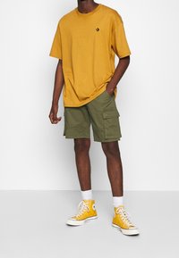 T-shirt en coton jaune à manches courtes, associée à un short cargo vert olive. La tenue est complétée par des baskets montantes jaunes.