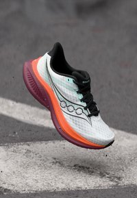 Λευκό και πορτοκαλί παπούτσι τρεξίματος Saucony με μαύρα κορδόνια, κρεμασμένο πάνω από ασφάλτινο δρόμο με λευκή γραμμή ζωγραφισμένη.