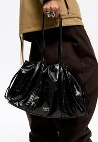 Czarna, błyszcząca torebka z marszczonym wzorem i długimi uchwytami, z wytłoczonym logo "BIMBA Y LOLA" na dole. Wykonana z fakturowanego materiału.