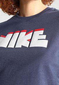 T-paita, joka on syvänsinistä puuvillaa ja jossa on rohkea valkoinen ja punainen "NIKE" -logo. Pyöreä kaula-aukko, lyhyet hihansuut ja sileä pinta.