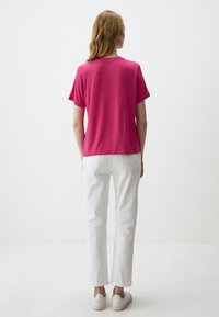 Korte mouwen, fuchsia top gecombineerd met witte jeans. Model gezien van achteren, met lichtgekleurde sneakers op een neutrale achtergrond.