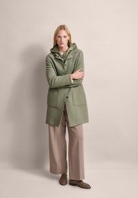 Grüne Wolljacke mit Kapuze, versehen mit Knopfverschluss und Vordertaschen. Getragen über beigen, weit geschnittenen Hosen und braunen Slippern. Neutrale Hintergrund.