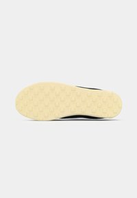 Μαύρη σόλα αθλητικού παπουτσιού με υφή, με κίτρινη καουτσούκ βάση που διαθέτει μικρά τετράγωνα για πρόσφυση. Λείες γωνίες και ελαφρύ σχέδιο.