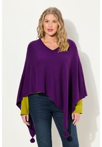 Poncho lavorato a maglia viola con scollo a V, bordi appuntiti e nappe decorative. Indossato sopra una maglietta a maniche lunghe verde e abbinato a jeans blu.
