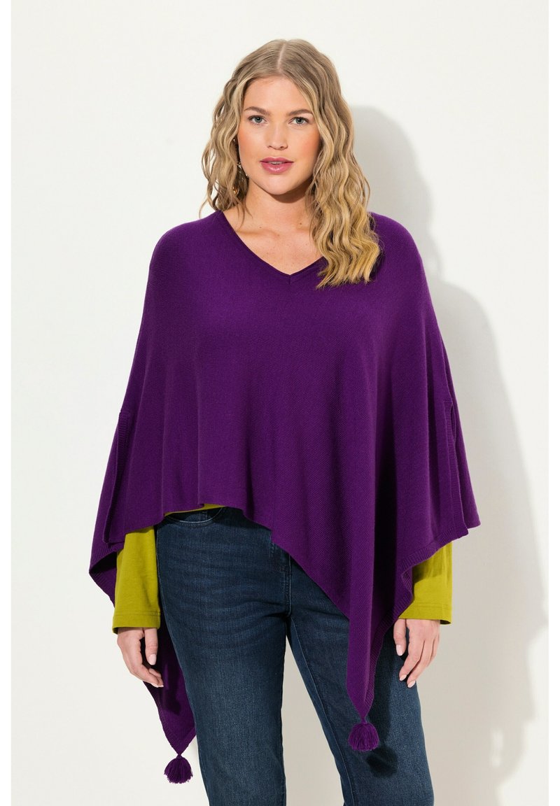 Poncho lavorato a maglia viola con scollo a V, bordi appuntiti e nappe decorative. Indossato sopra una maglietta a maniche lunghe verde e abbinato a jeans blu.