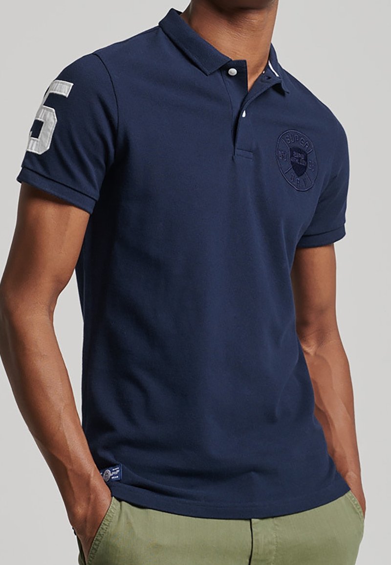 Homme portant un polo à manches courtes bleu marine avec le chiffre blanc 6 sur la manche et un pantalon vert olive, les mains dans les poches.