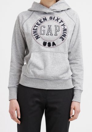 Jasnoszara bluza z kapturem z przednią kieszenią z okrągłym logo "NINETEEN SIXTY-NINE GAP USA", noszona z czarnymi spodniami.