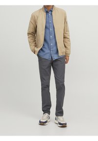 Beige Bomberjacke über einem blauen Jeanshemd, graue Hose und Turnschuhe mit blauen und cremefarbenen Akzenten. Lässiges, strukturiertes Design mit einem Reißverschlusstasche.