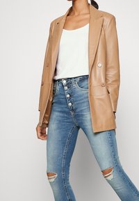 Tan läderblazer med dubbla knappar, bärs över en vit tanktop. Kombinerad med blå smala jeans med slitna knädetaljer och knappstängning.