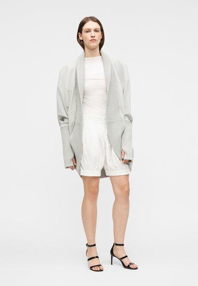 Γυναίκα που φοράει ανοιχτό γκρι φαρδιά ζακέτα, λευκό διάφανο τοπ, άσπρα ελαστικά σορτς και μαύρα πέδιλα με λουράκια και ψηλό τακούνι, στέκεται μπροστά σε απλό φόντο.